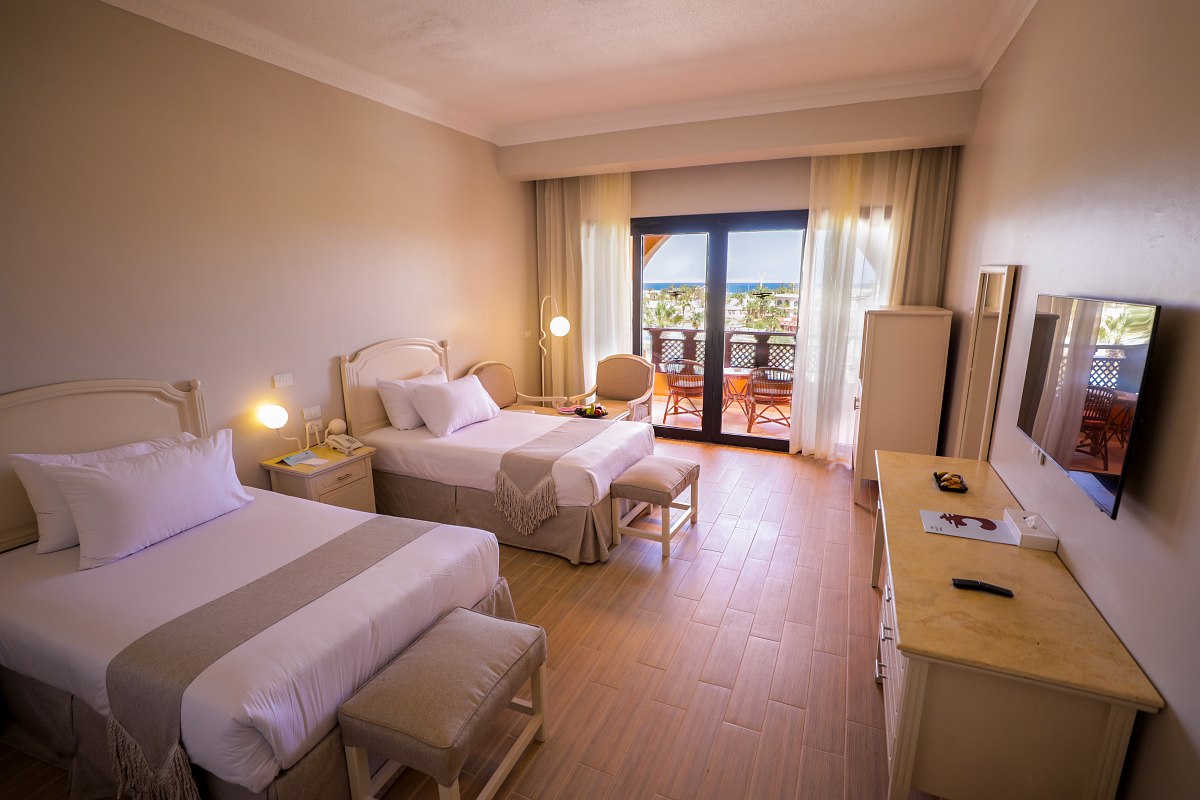 Hell eingerichtetes Hotelzimmer mit Doppelbett, zwei Hockern, Sesseln und Terrasse mit Blick auf die Natur.