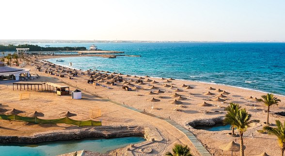 Weitläufiger, sonniger Sandstrand mit vielen Sonnenschirmen, klarem blauem Meer und Palmen am Ufer.