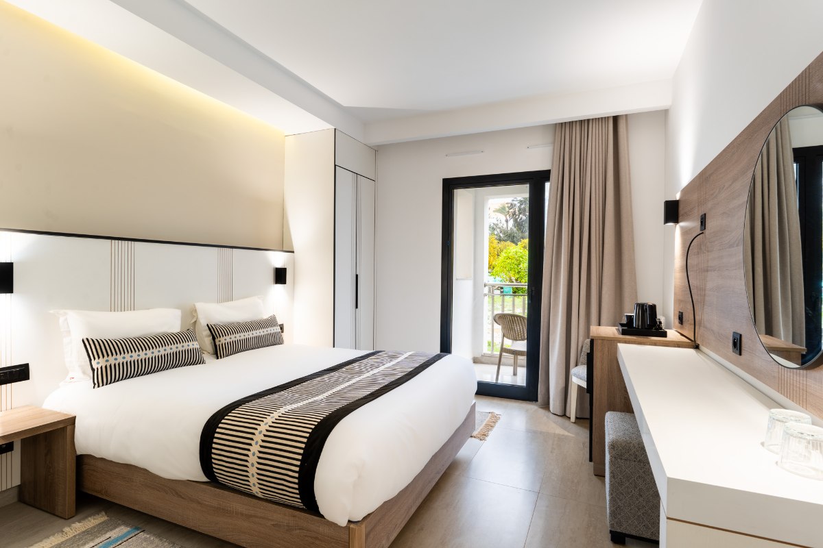 Hell eingerichtetes Hotelzimmer mit zwei Einzelbetten, blauem Sofa, großem Fenster und floralem Wandbild.