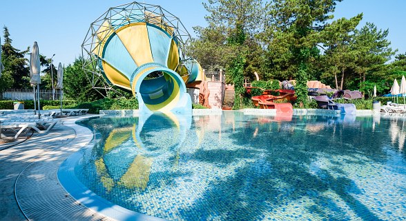 Große gelb-blaue Wasserrutsche, die in einen klaren Pool mit Sonnenliegen am Rand mündet, umgeben von grüner Bepflanzung.