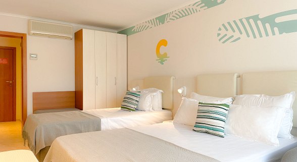 Doppelzimmer mit zwei Betten, beige Bettwäsche, weißen Wänden und Holzkleiderschrank, mit gemusterten Kissen und Wanddekor.