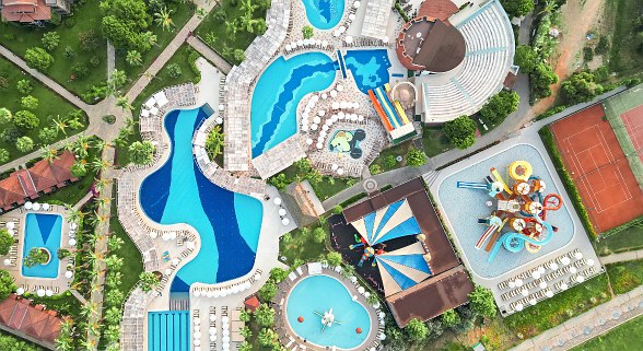 Luftaufnahme einer großen Hotelanlage mit mehreren Pools, Wasserrutschen, Amphitheater und Tennisplatz, umgeben von viel Grün.
