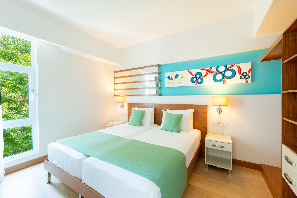 Hell eingerichtetes Zimmer mit zwei Einzelbetten, grünen Kissen, Nachttisch, offener Garderobe und farbenfroher Wanddekoration.