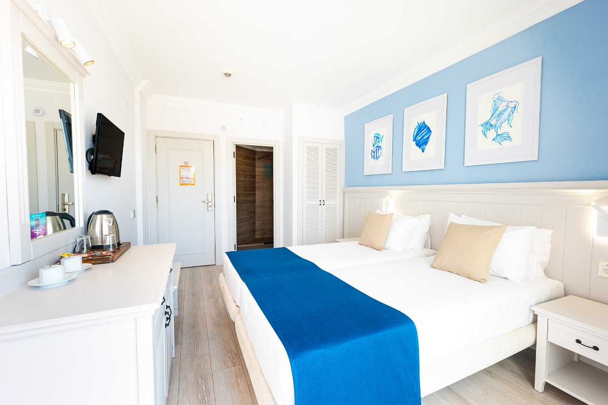 Helles Zimmer mit zwei Betten, weißen Wänden und blauen Akzenten. Kleiner Schreibtisch mit Spiegel, Fernseher an der Wand, Koffer auf der Bank. Sauber und modern gestaltet.