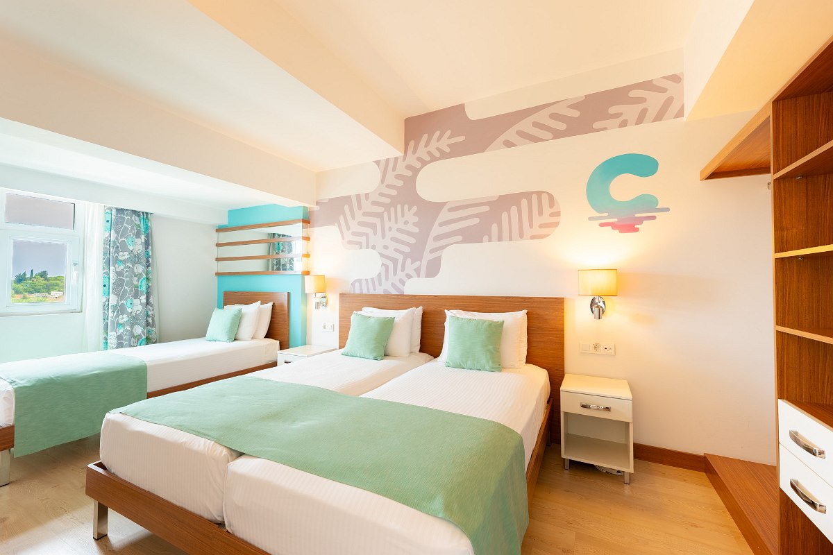 Gemütliches Apartment mit Küchenzeile, Sofa und kleinem Esstisch. Durch die offene Tür sieht man ein Schlafzimmer mit grünem Bett und einem kleinen TV. Balkon mit Sitzmöbeln im Hintergrund.