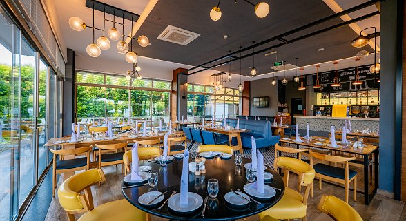 Helles Restaurant mit Holz- und Metallstühlen, Holztischen, runden Platzsets und großen Fenstern mit Blick auf grüne Bäume.
