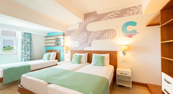 Modernes Doppelzimmer mit hellen Wänden, zwei Einzelbetten, blauem Bettläufer und Zugang zu einem Balkon mit Vorhängen.