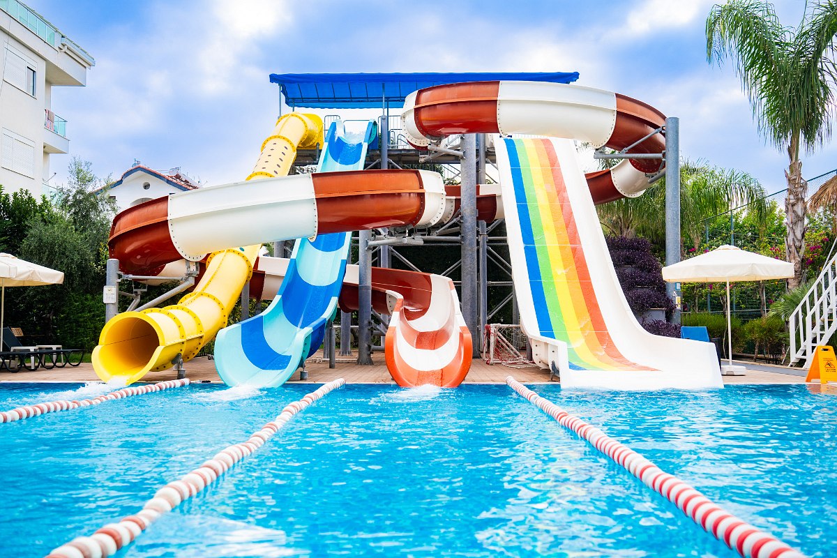 Pool mit bunten Wasserrutschen und blauen Liegestühlen am Beckenrand, daneben ein modernes, helles Hotelgebäude.