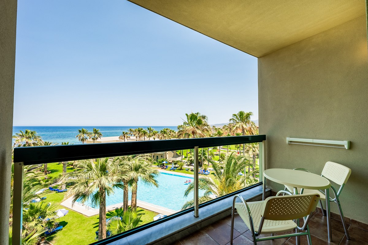 Balkon mit Tisch und Stühlen, Blick auf Poolanlage, Palmen und das Meer unter klarem, blauem Himmel.