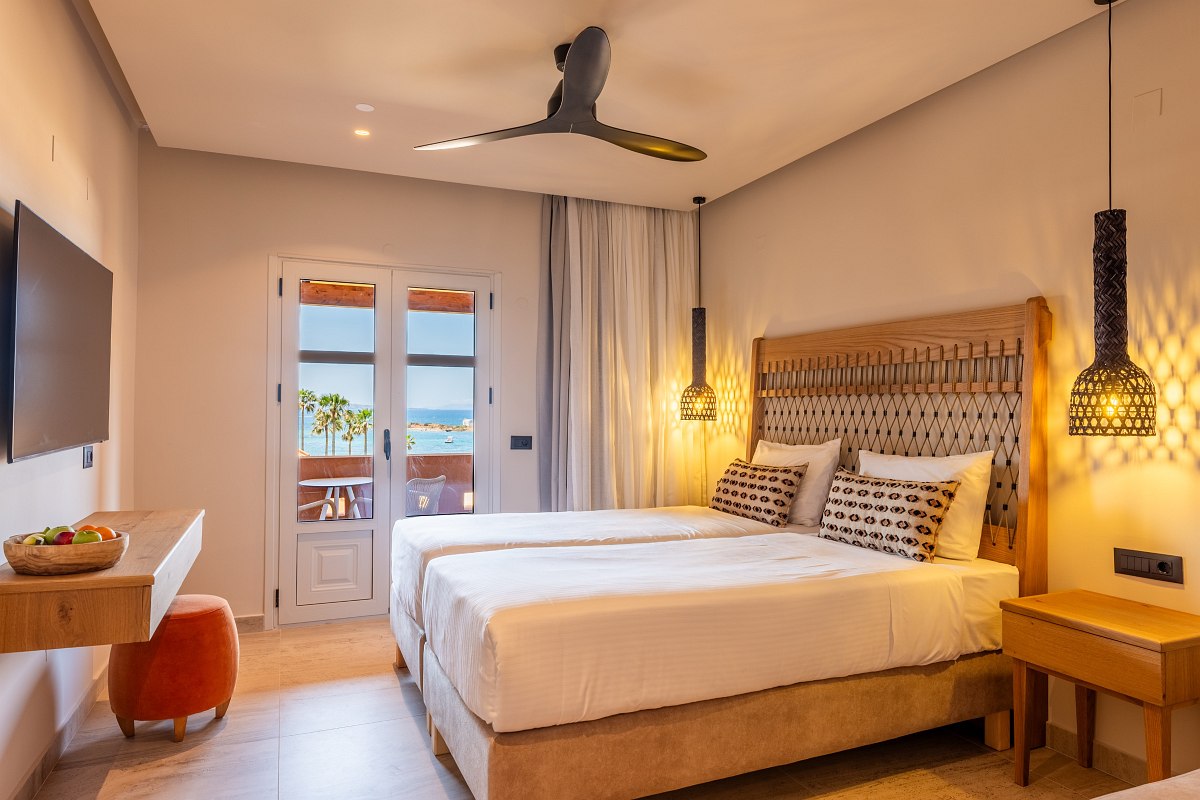Helles Hotelzimmer mit Doppelbett, gemütlicher Beleuchtung, TV und Balkon mit Meerblick und Palmen.