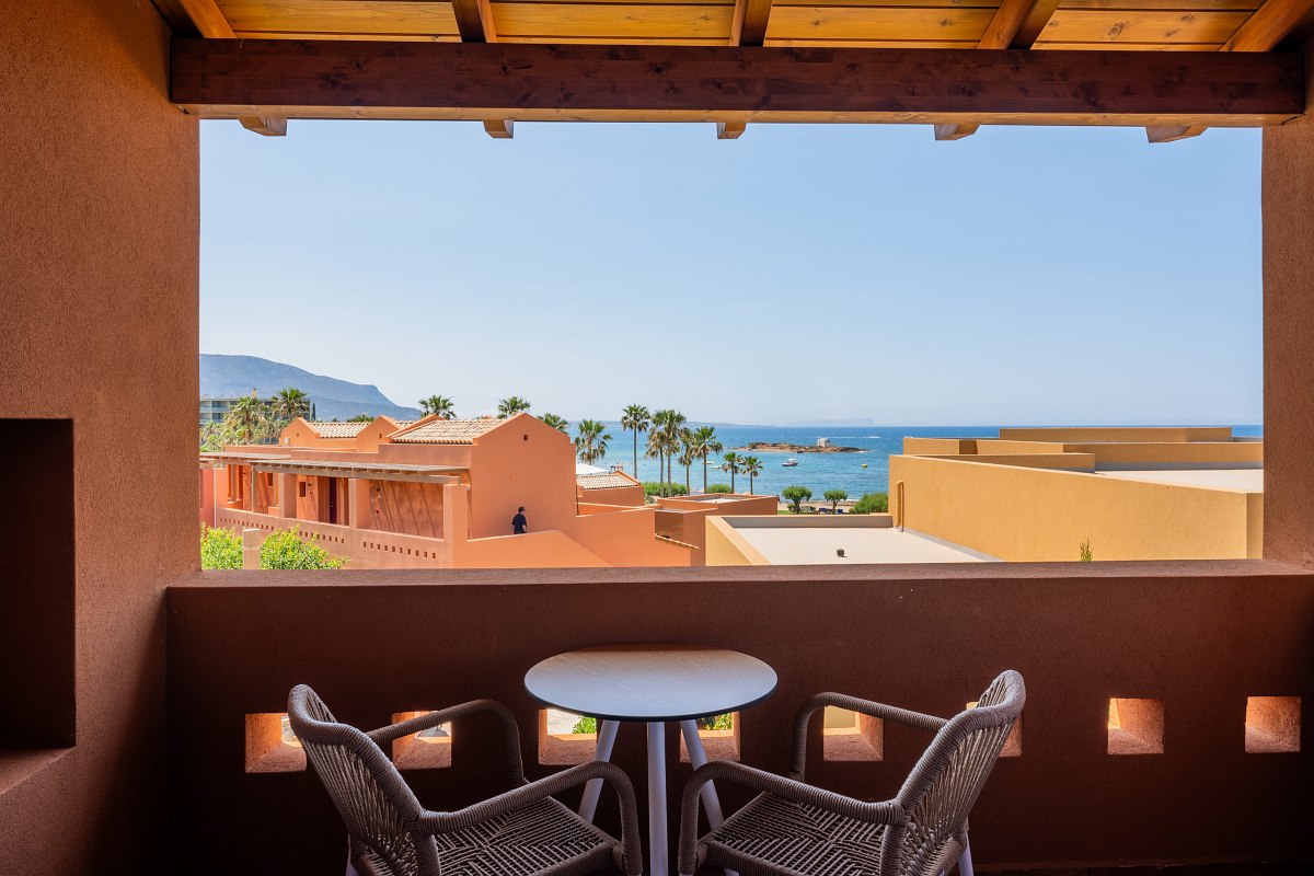 Balkon mit zwei Stühlen und Tisch, Blick auf mediterrane Gebäude, Palmen und das blaue Meer mit Inseln im Hintergrund.