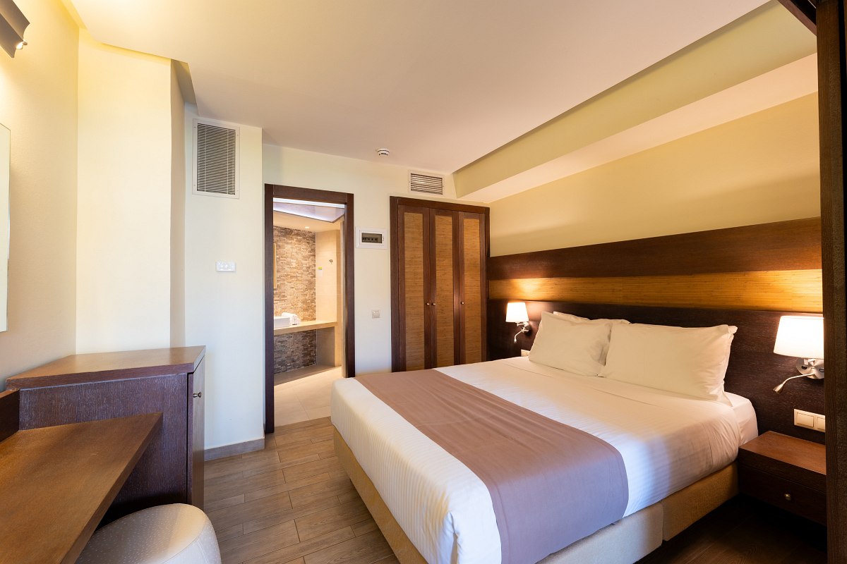 Helles Hotelzimmer mit Doppelbett, Holzdetails, Schreibtisch und Blick ins moderne Badezimmer mit Natursteinwand.