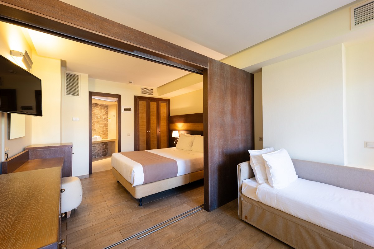 Helles Hotelzimmer mit Doppelbett, Sofa-Bett, Holzdetails, Schreibtisch und Zugang zum offenen Badezimmerbereich.