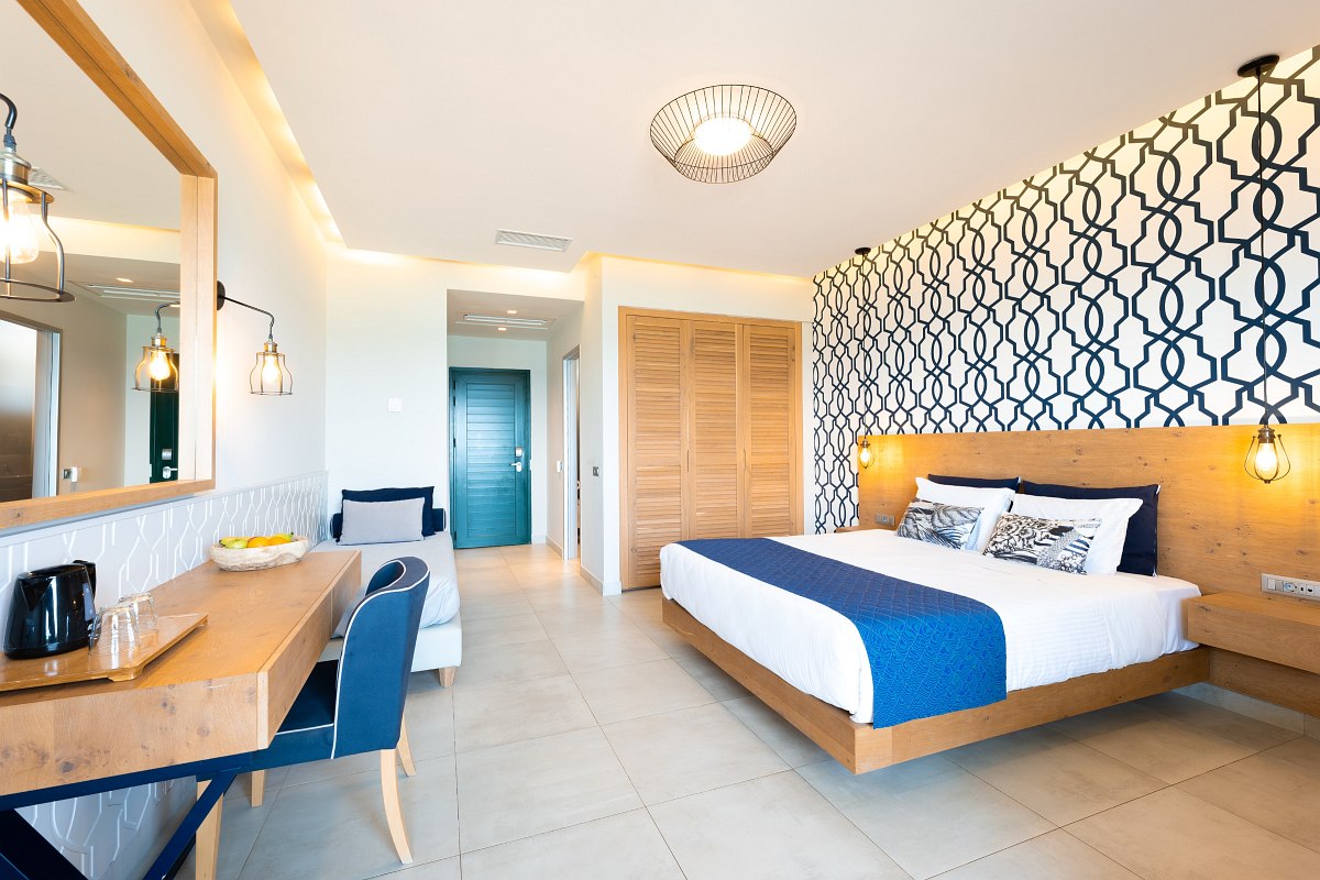 Helles Hotelzimmer mit Doppelbett, gemusterten Wänden, Sofa, Schreibtisch, Holzschrank und türkisfarbener Tür.