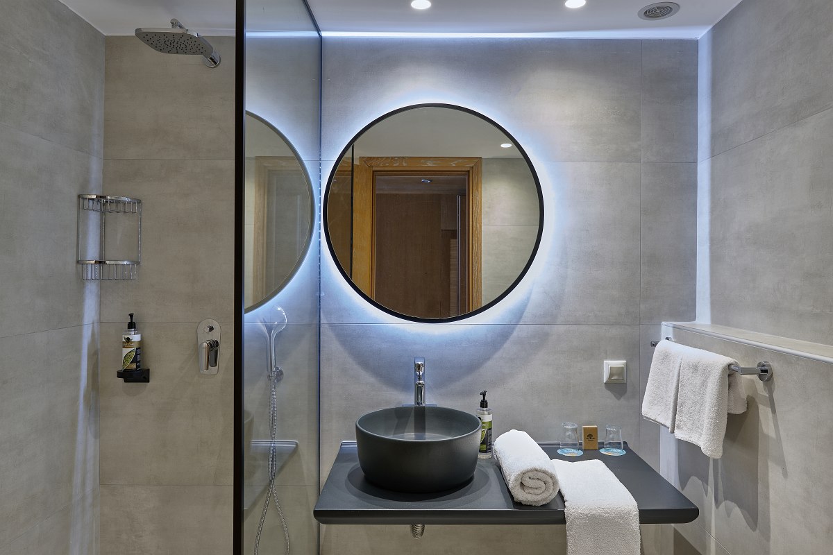 Modernes Badezimmer mit rundem, beleuchtetem Spiegel, schwarzem Waschbecken und begehbarer Dusche.