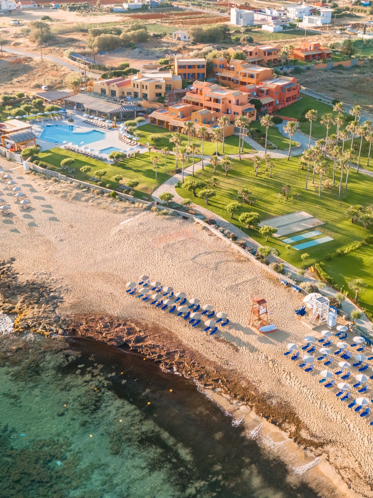 Luftaufnahme eines Strandresorts mit Pool, Liegestühlen, Sonnenschirmen und Palmen direkt am Meer bei sonnigem Wetter.