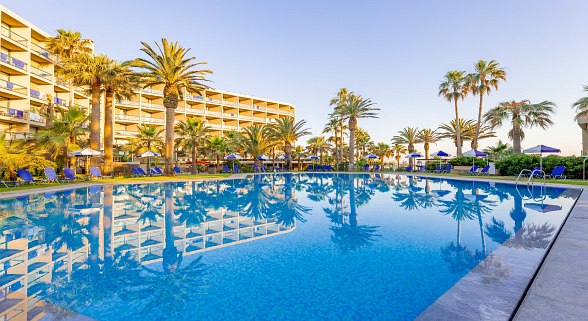 Großer Hotelpool mit Palmen und Sonnenliegen, im Hintergrund mehrstöckiges Hotelgebäude bei klarem Himmel.
