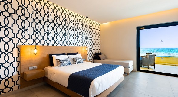 Helles Hotelzimmer mit Doppelbett, gemusterten Wänden, Sofa, Schreibtisch, Holzschrank und türkisfarbener Tür.