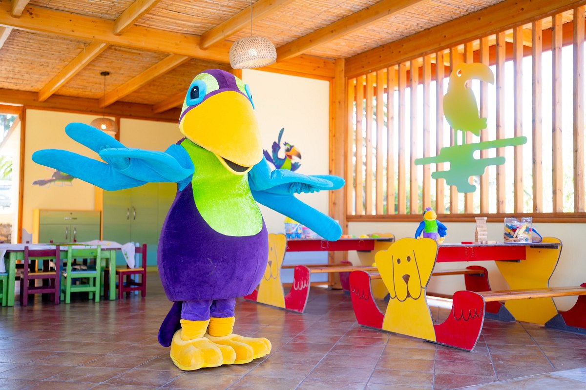 Bunter Maskottchen-Vogel in einem farbenfrohen Kinderbereich mit Holzbänken, Tischen und buntem Spielzeug.