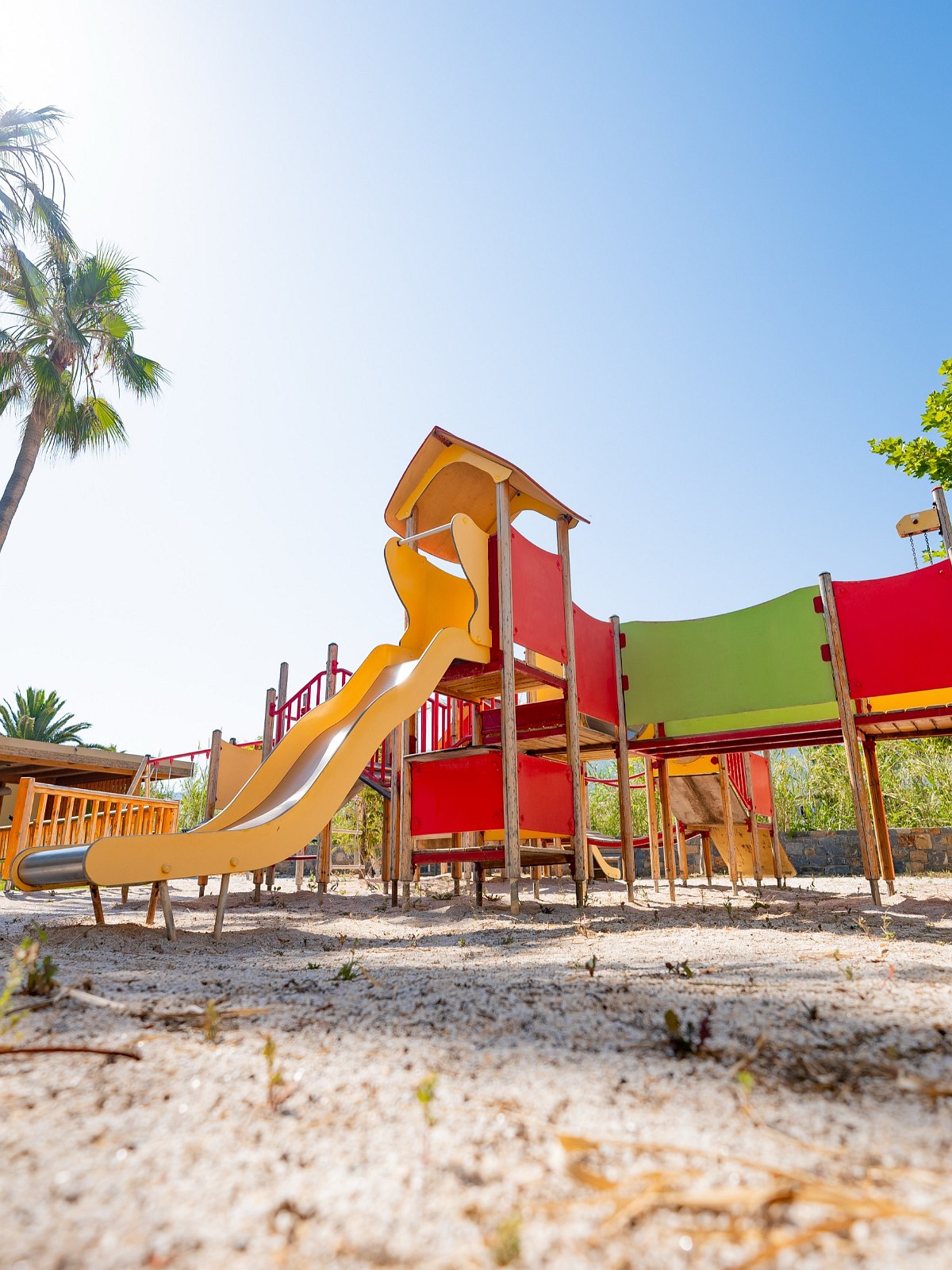 Bunter Kinderspielplatz mit Rutsche und Kletterwand im Sandbereich, umgeben von Palmen und Bäumen bei sonnigem Wetter.