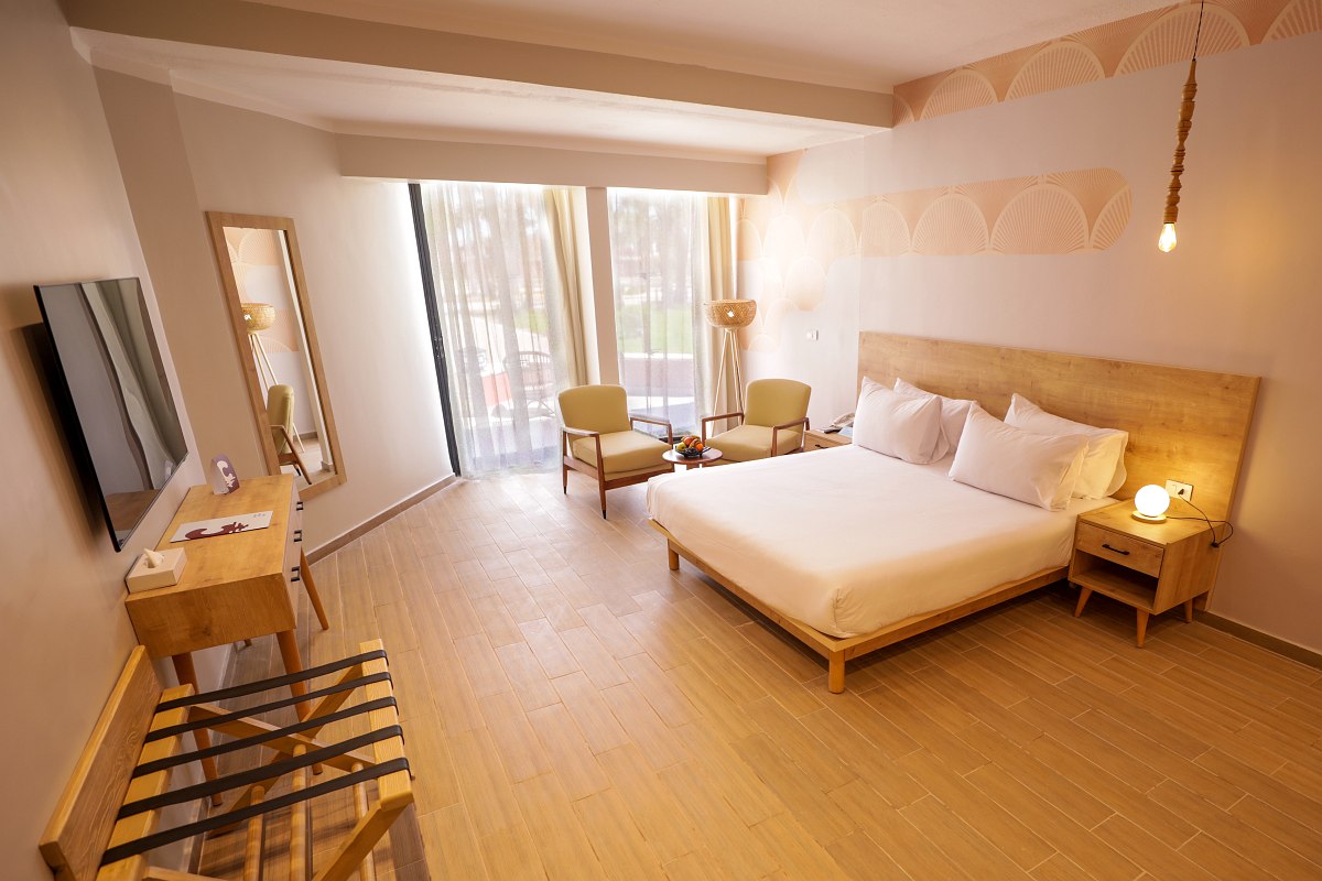 A bright hotel room with two large beds, ornate headboards, a small seating area, and a balcony with outdoor furniture visible through sliding glass doors.