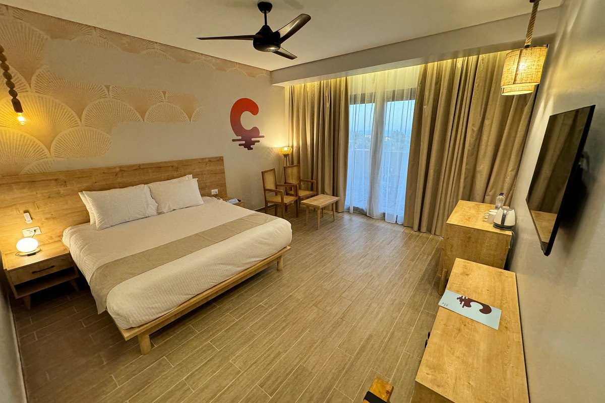 A clean and bright hotel room with a large bed, two cushioned stools at the foot, simple artwork on the wall, a small table with a tray of food, and soft lighting creating a cozy atmosphere.