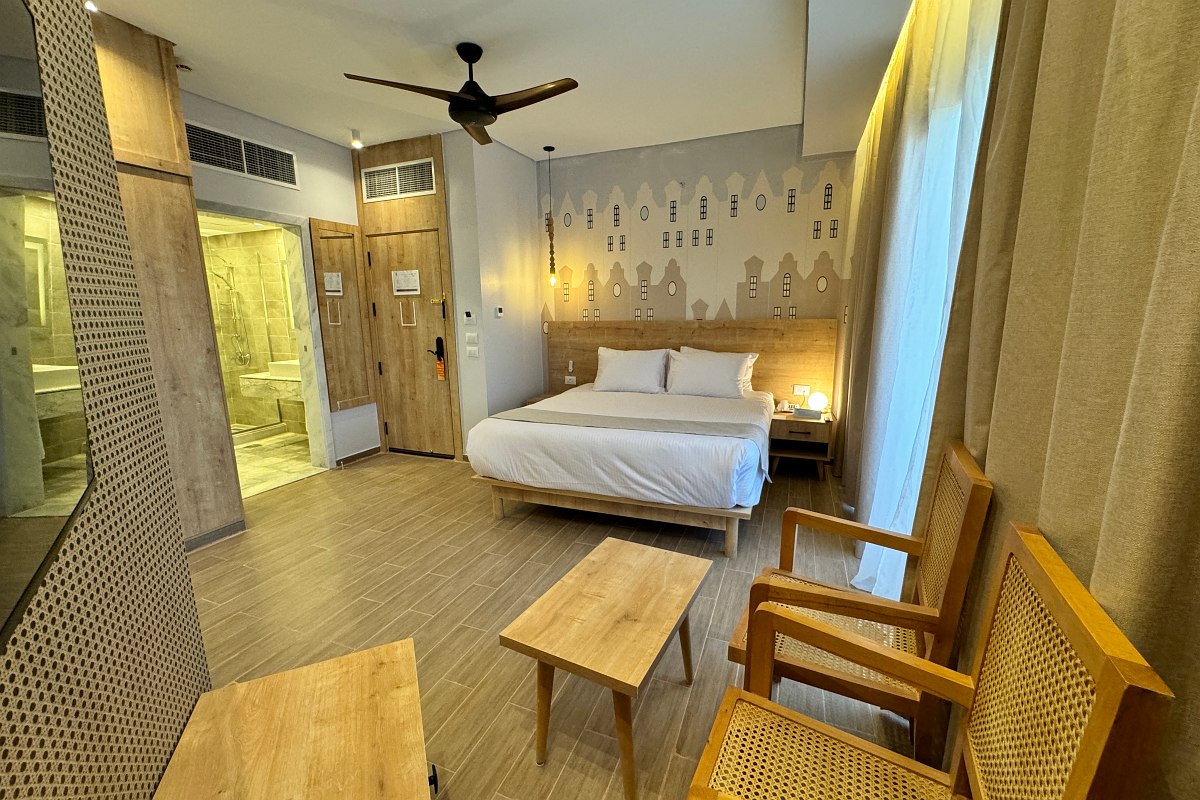 A cozy hotel room with two beds, classic white furniture, and a balcony with outdoor seating, letting in natural light through large sliding doors.