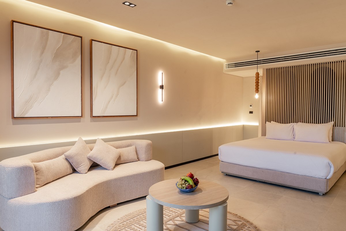 A bright room with two single beds, matching benches, an armchair with a small table holding snacks, and simple wall art.