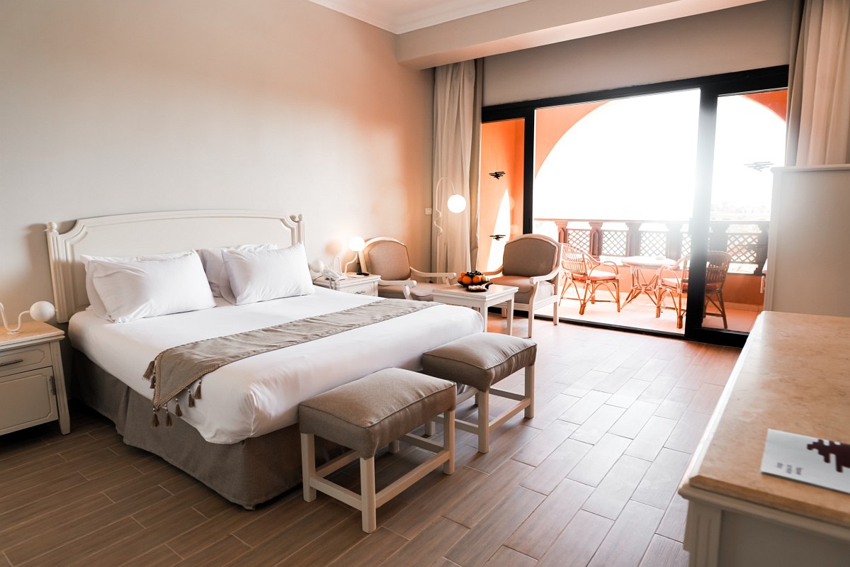 A modern hotel room with a large, neatly made bed, two upholstered stools, cozy chairs, and a balcony with outdoor seating overlooking a bright, airy view.