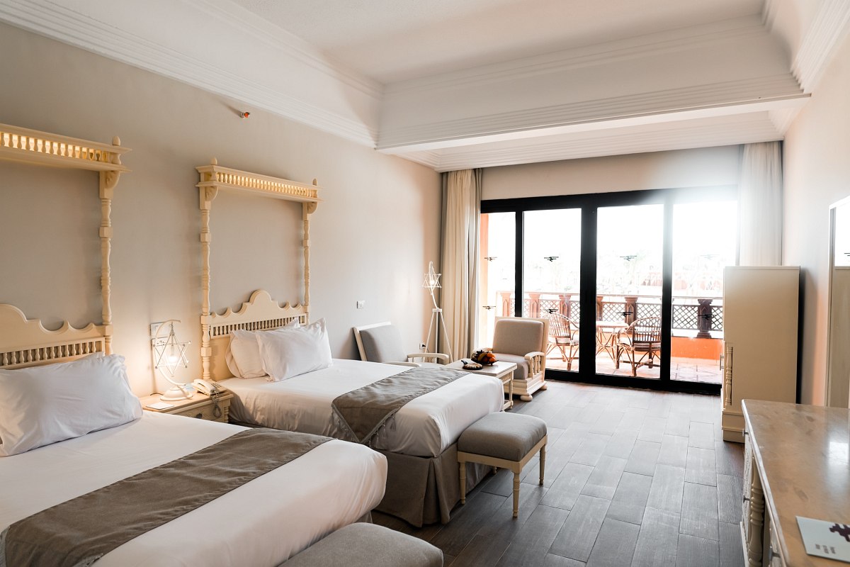 A bright hotel room with two large beds, ornate headboards, a small seating area, and a balcony with outdoor furniture visible through sliding glass doors.