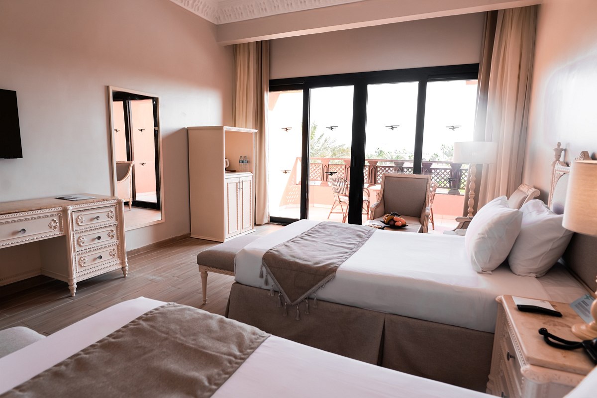A cozy hotel room with two beds, classic white furniture, and a balcony with outdoor seating, letting in natural light through large sliding doors.
