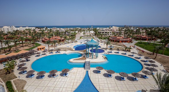A large resort pool area with a central gazebo, surrounded by numerous sun loungers and umbrellas, palm trees, and buildings with a sea view in the background.