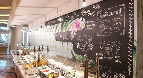 A well-lit buffet area with a variety of dishes displayed under a white canopy, featuring a decorative blackboard wall with restaurant-themed chalk art and greetings.