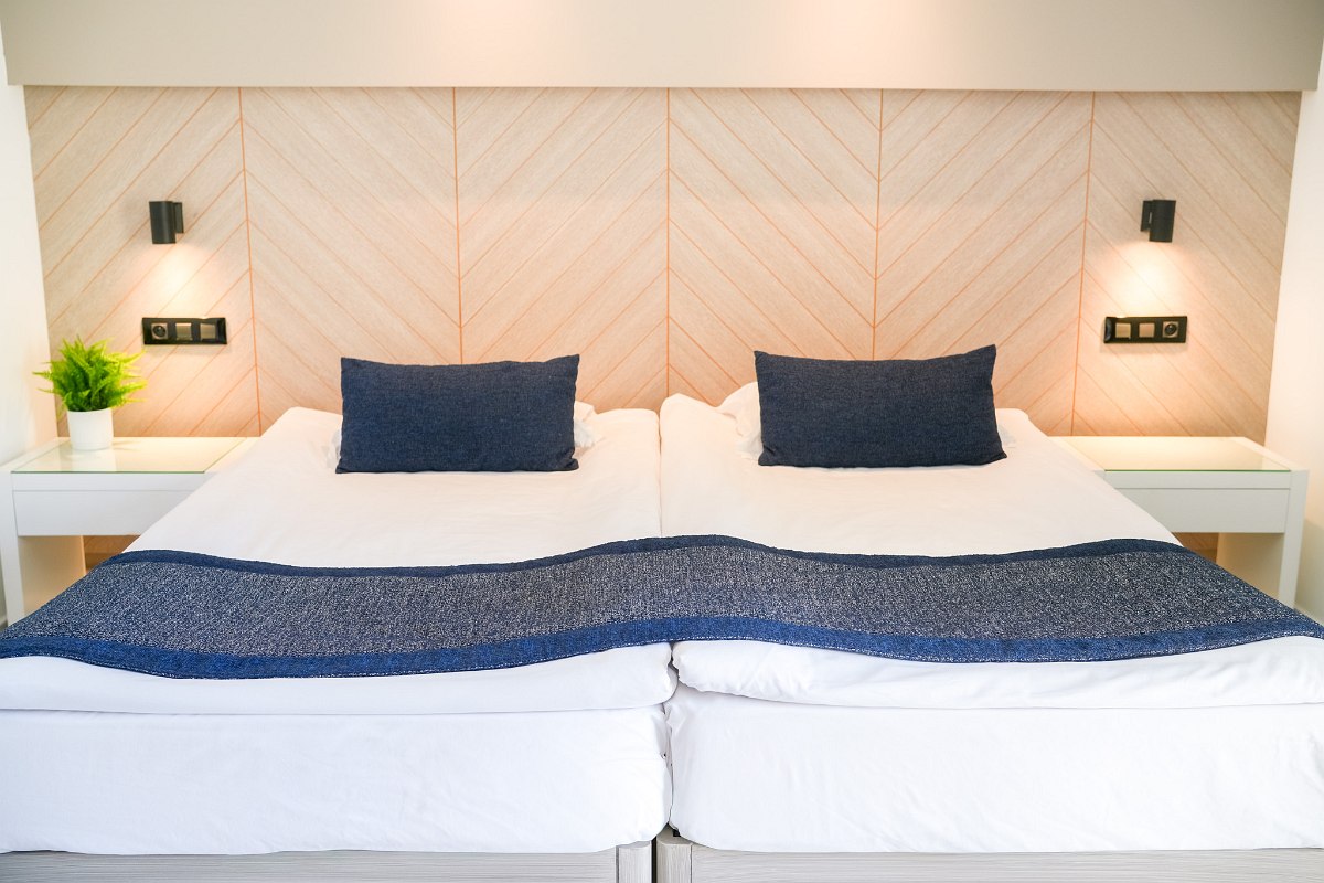 A modern hotel room with a large bed, patterned bedding, a small desk with chairs, a wall-mounted TV, and a balcony with outdoor seating.