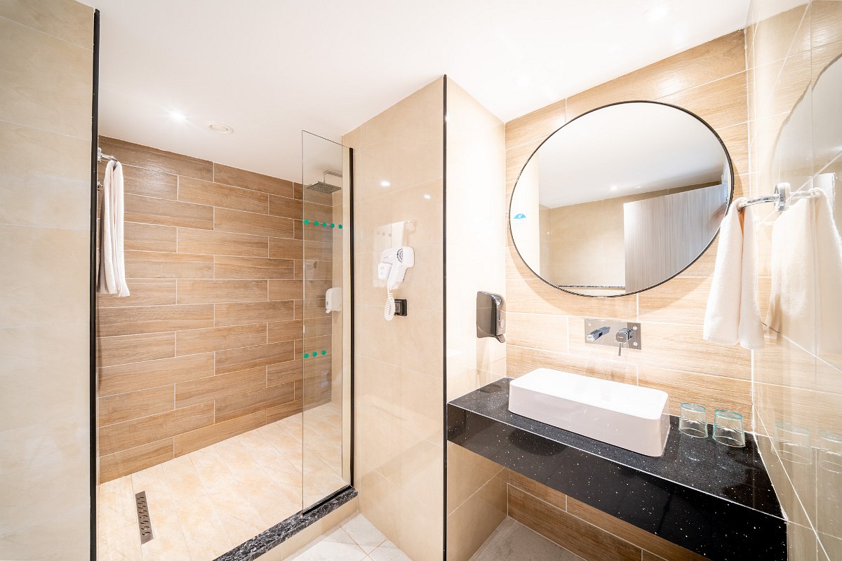 A sleek modern bathroom featuring a black vanity with a white vessel sink, a large rectangular mirror, a walk-in glass shower, and neutral beige tiles.