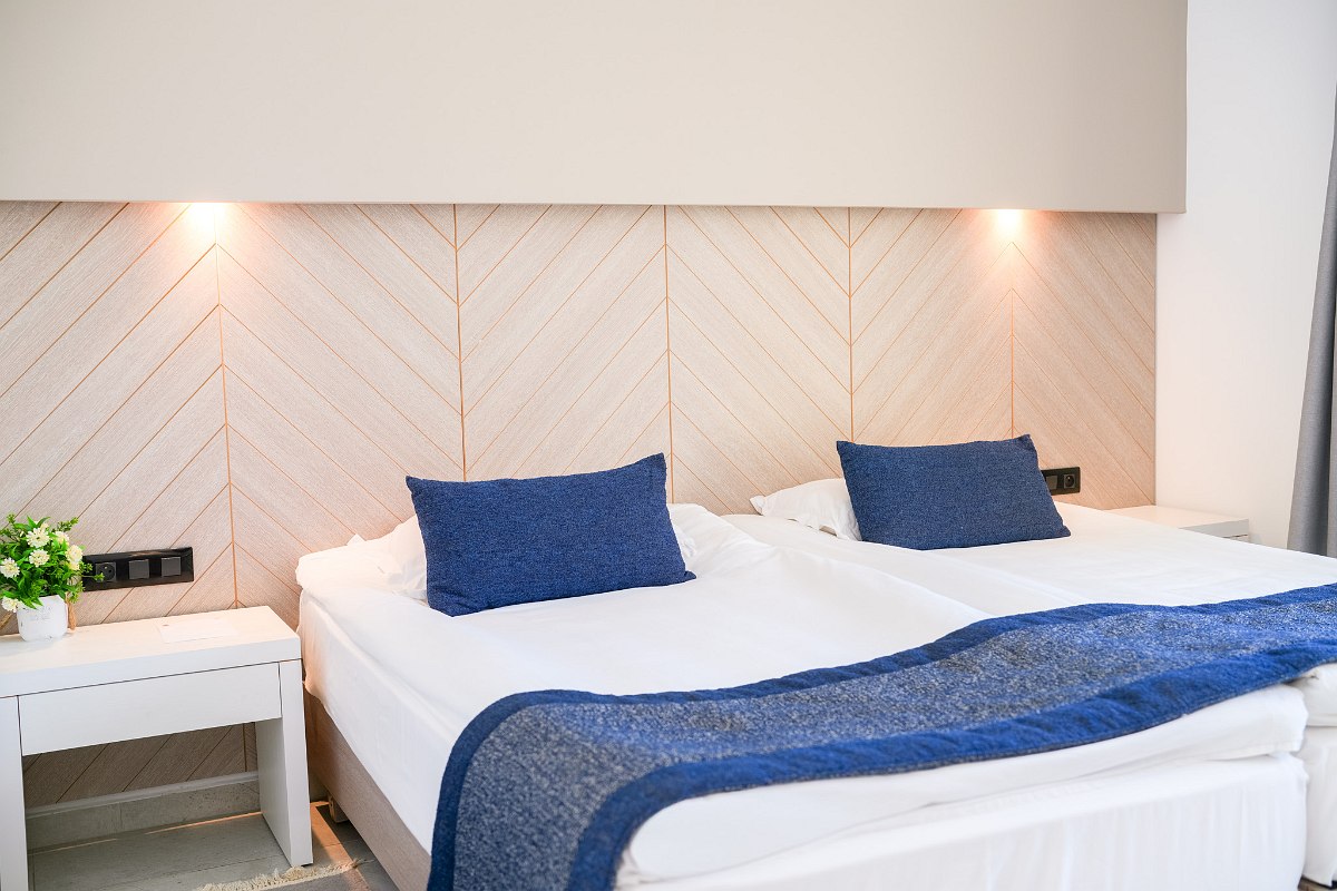 A cozy hotel room with twin beds, green accents, a wooden desk and chair, wall lamps, and a sliding glass door opening to a small patio with garden views.