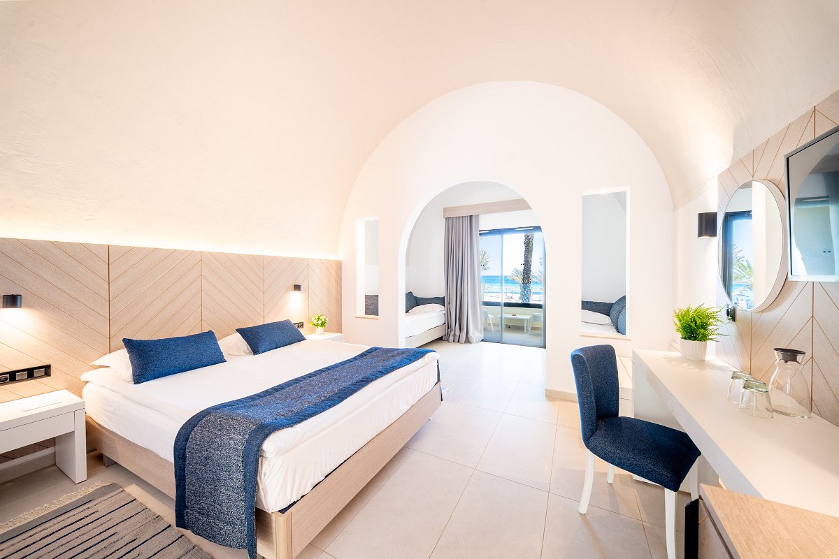 A bright hotel room with three single beds, green bed runners and pillows, a wooden bench, and a window with yellow and green curtains overlooking palm trees.