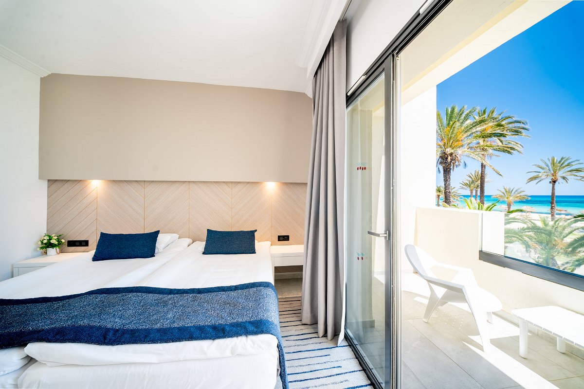 A spacious hotel room with a vaulted ceiling, a large bed with green bedding, wooden furniture, a TV, and a balcony with a sea view.