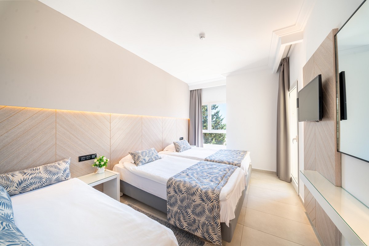 A modern hotel room with a large bed, a seating area with two chairs and a desk, a wall-mounted TV, and a sliding glass door opening to a balcony. Another bedroom is visible through an open door.