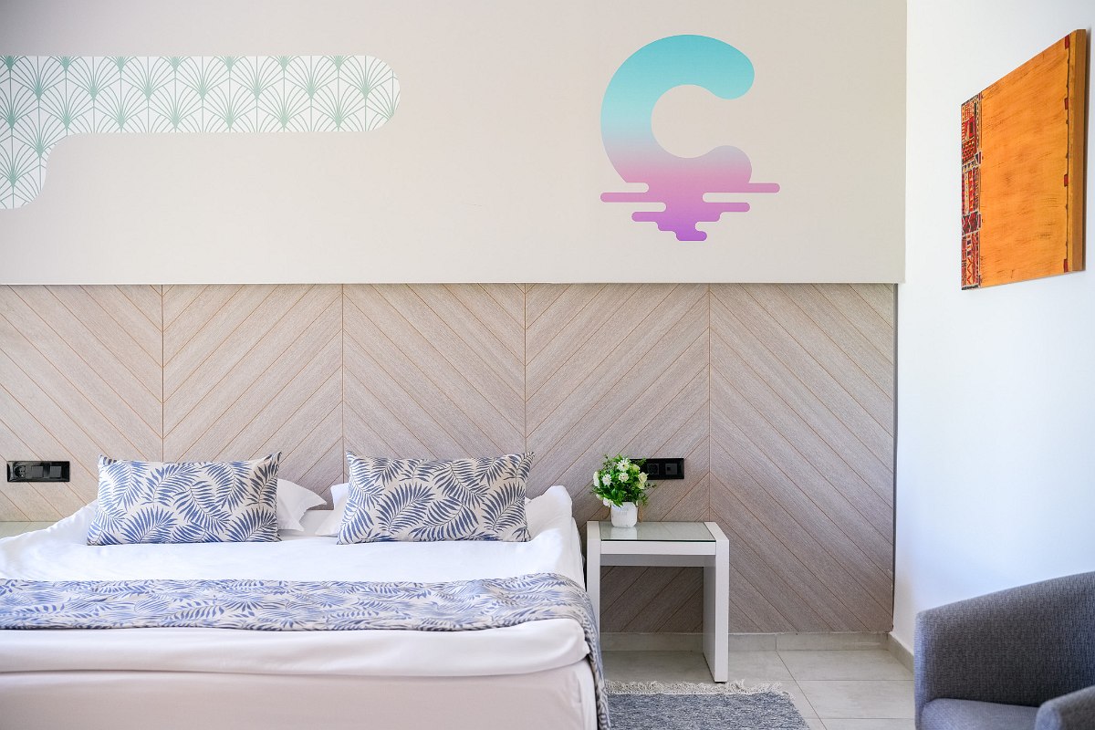 A modern hotel room with a large bed, gray and beige bedding, a decorative wall with soft patterns and a logo, a window with curtains, and a clean, minimalistic design.