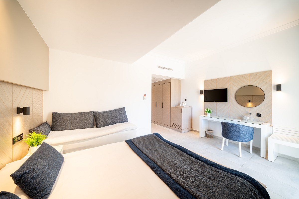 A modern hotel room with a large bed, gray and beige bedding, a decorative wall with soft patterns and a logo, a window with curtains, and a clean, minimalistic design.