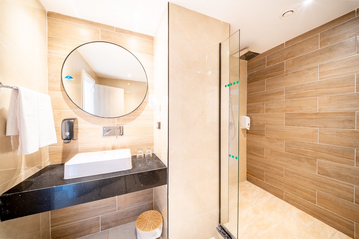 A modern bathroom with beige tiles, a glass shower enclosure, a black vanity with a white vessel sink, a round mirror, and a toilet partially visible behind a wall. The shower glass features the text 