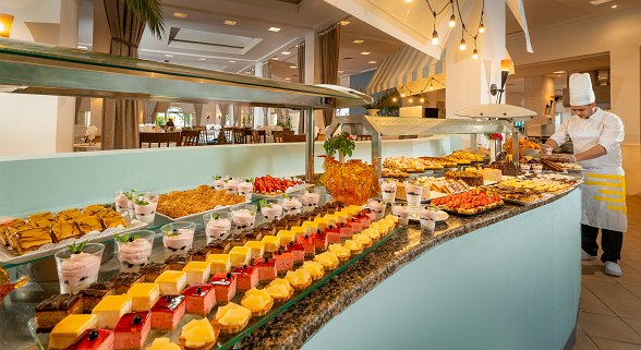 A dessert buffet with a variety of colorful cakes, pastries, and parfaits displayed on a long curved counter, with a chef arranging dishes in the background.
