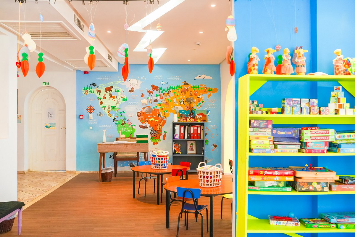 A colorful kids’ playroom with tables, toys, games, a large world map mural on the wall, and children engaged in face painting and crafts activities.