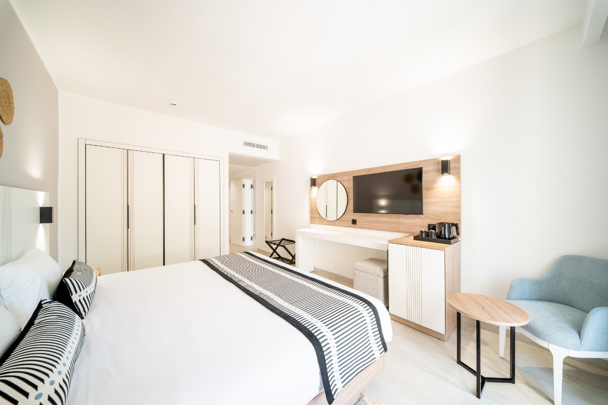 A bright hotel room with a large bed, blue and beige accents, patterned rugs, a balcony with outdoor seating, and a decorative blue wall.