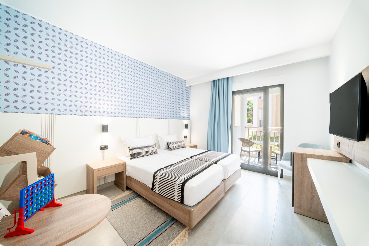 A bright hotel room with three single beds, blue bedding accents, a playful teepee, a patterned blue wall, and a balcony with outdoor seating.