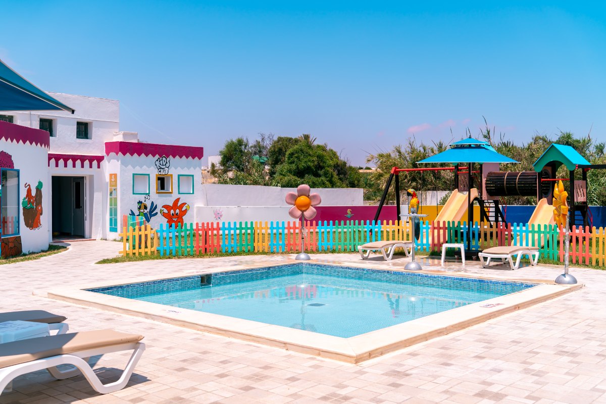 A colorful spiral water slide in a pool area, designed with a yellow base decorated with pastel hexagon patterns, surrounded by palm trees and fencing.