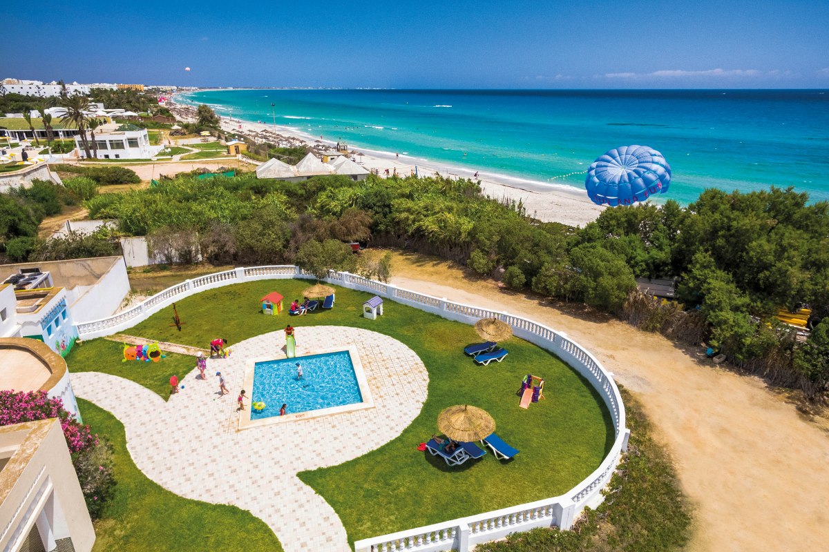 A colorful spiral water slide in a pool area, designed with a yellow base decorated with pastel hexagon patterns, surrounded by palm trees and fencing.