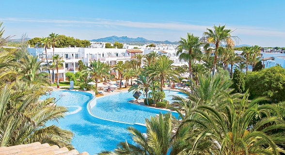 A large, winding resort pool surrounded by lush palm trees and white buildings under a clear blue sky.