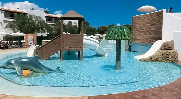 A playful kids’ water park area with shallow pools, slides, a dolphin sculpture, and a palm tree water feature, surrounded by resort buildings and sun loungers.